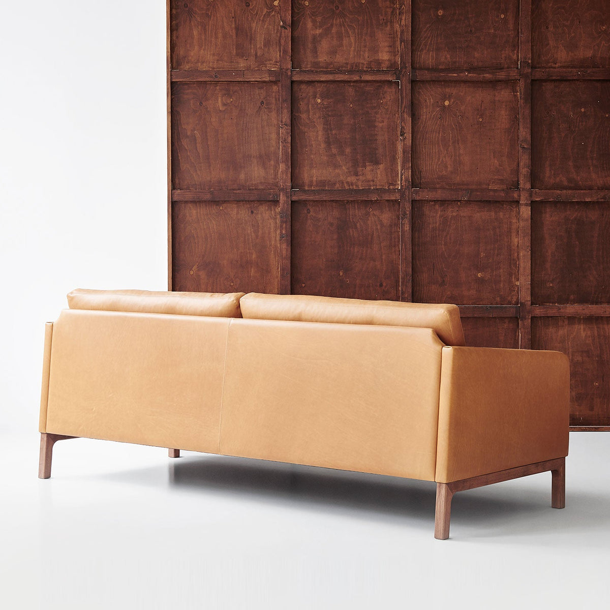 A modern tan leather sofa with wooden legs, positioned in front of a dark brown wood-paneled wall and next to a black tripod floor lamp.