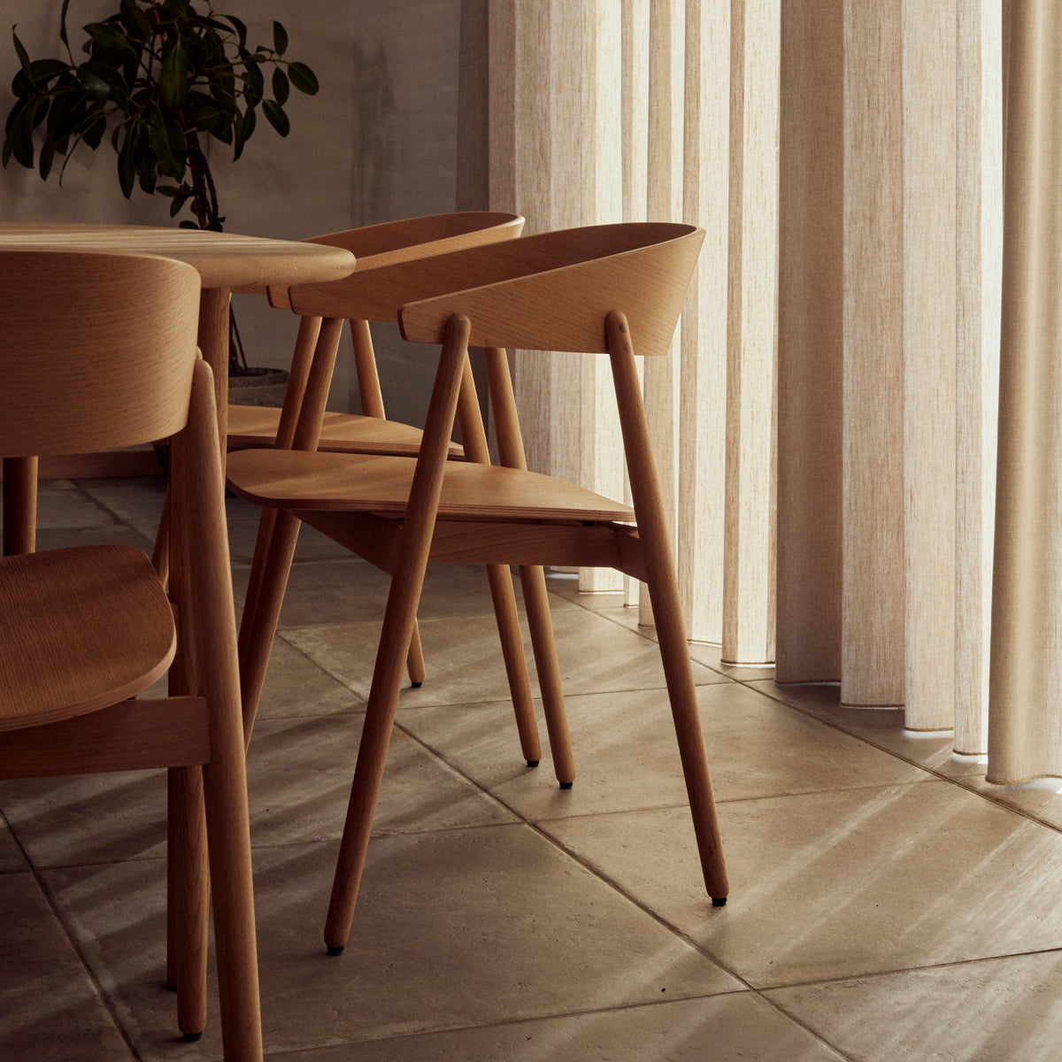 A dining area with light natural wood chairs and a matching table, positioned in front of sheer cream-colored curtains by a window. The room features warm neutral walls and tiled flooring, with a potted green plant in the background.