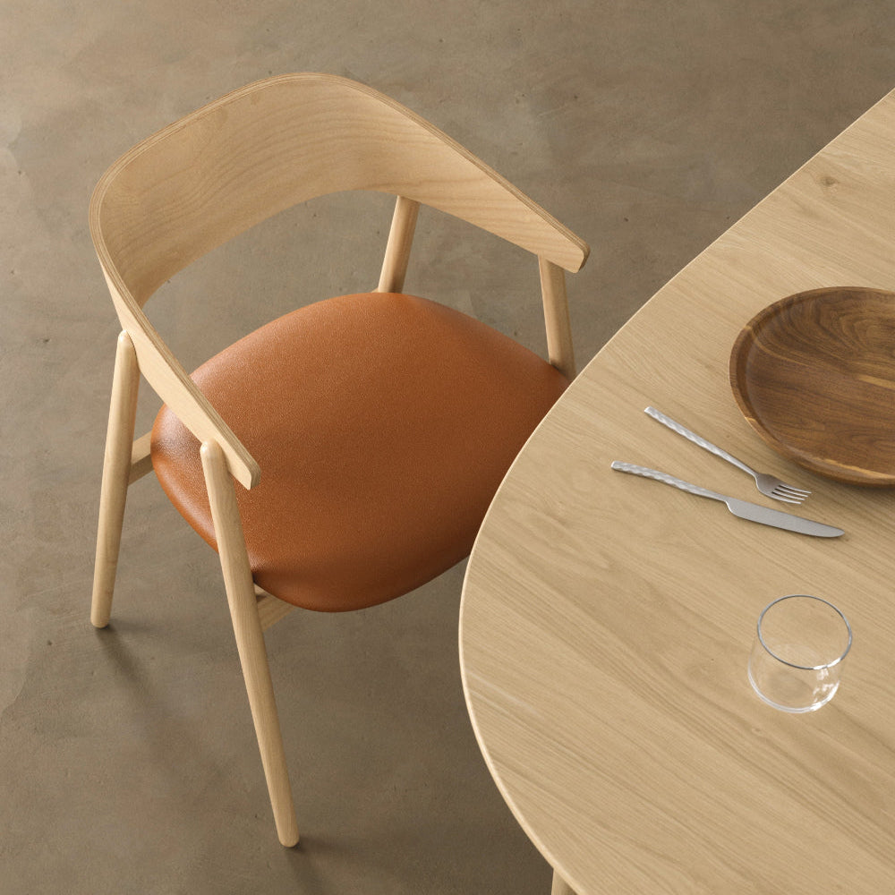 A modern dining chair with a light natural wood frame and a warm brown leather seat, positioned next to a matching light wood table on a concrete floor.