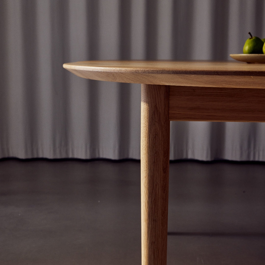 Close-up of a light brown wooden dining table showing the bevelled edge of the tabletop and the front leg, set against a dark gray floor and grey curtain backdrop.