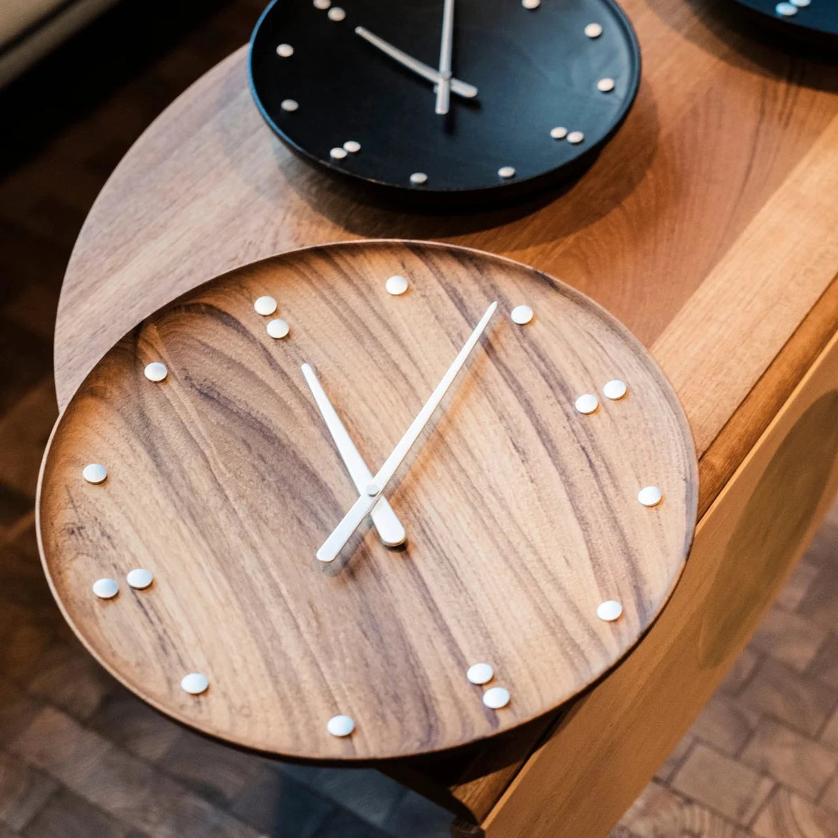 A round wooden wall clock with a natural teak wood grain face, silver hands, and small silver dot hour markers, shown mounted on a wooden table. A second black wall clock is partially visible behind it.