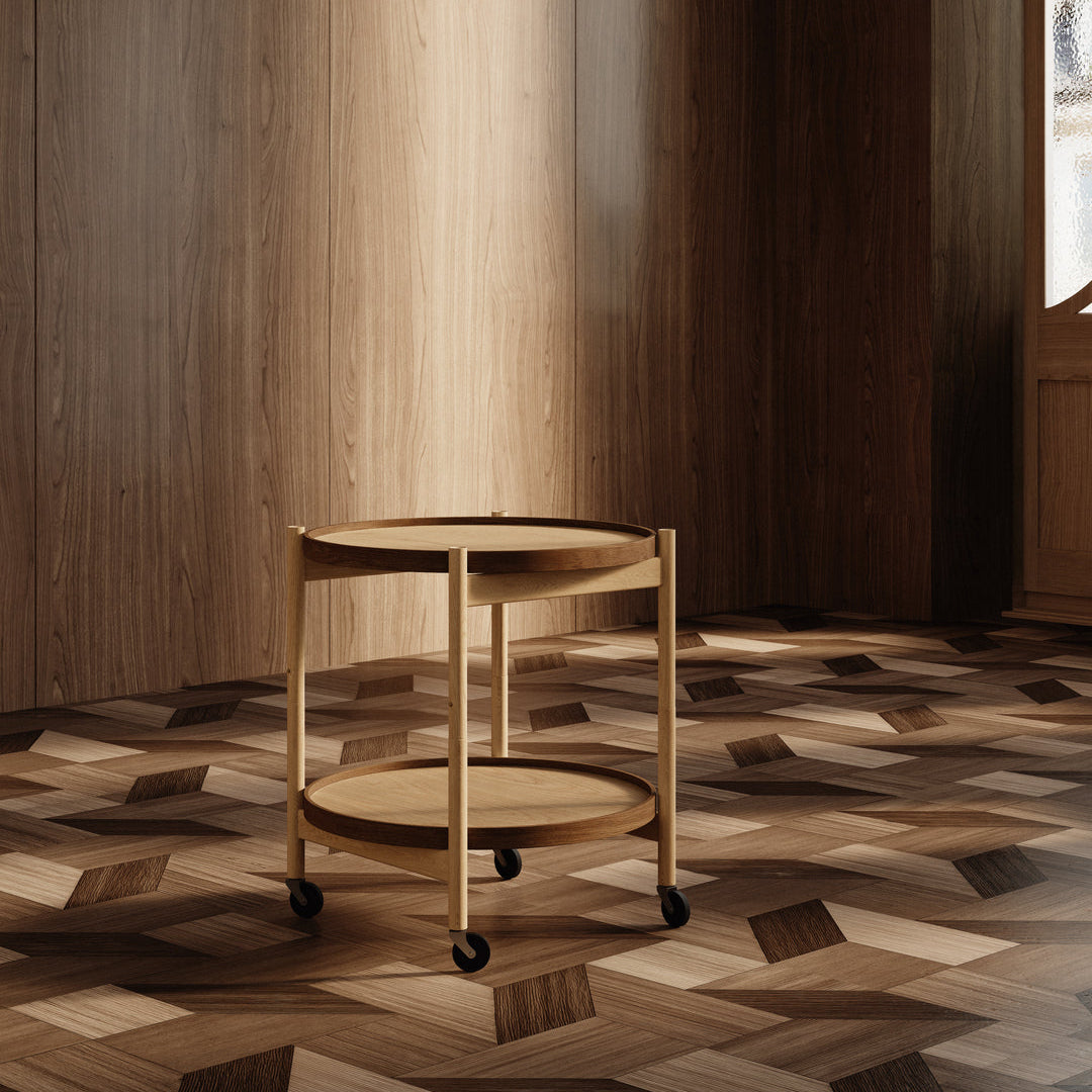 A small round wooden rolling side table with two shelves and black caster wheels, placed in a warm wood-paneled room with a geometric parquet floor.