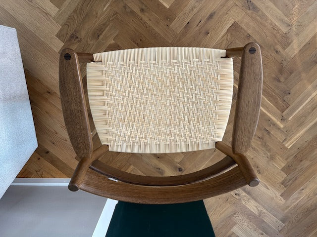 A wooden chair viewed from above, featuring a curved medium-brown wood frame and a woven natural-fiber seat. The chair is placed on a herringbone wood floor.