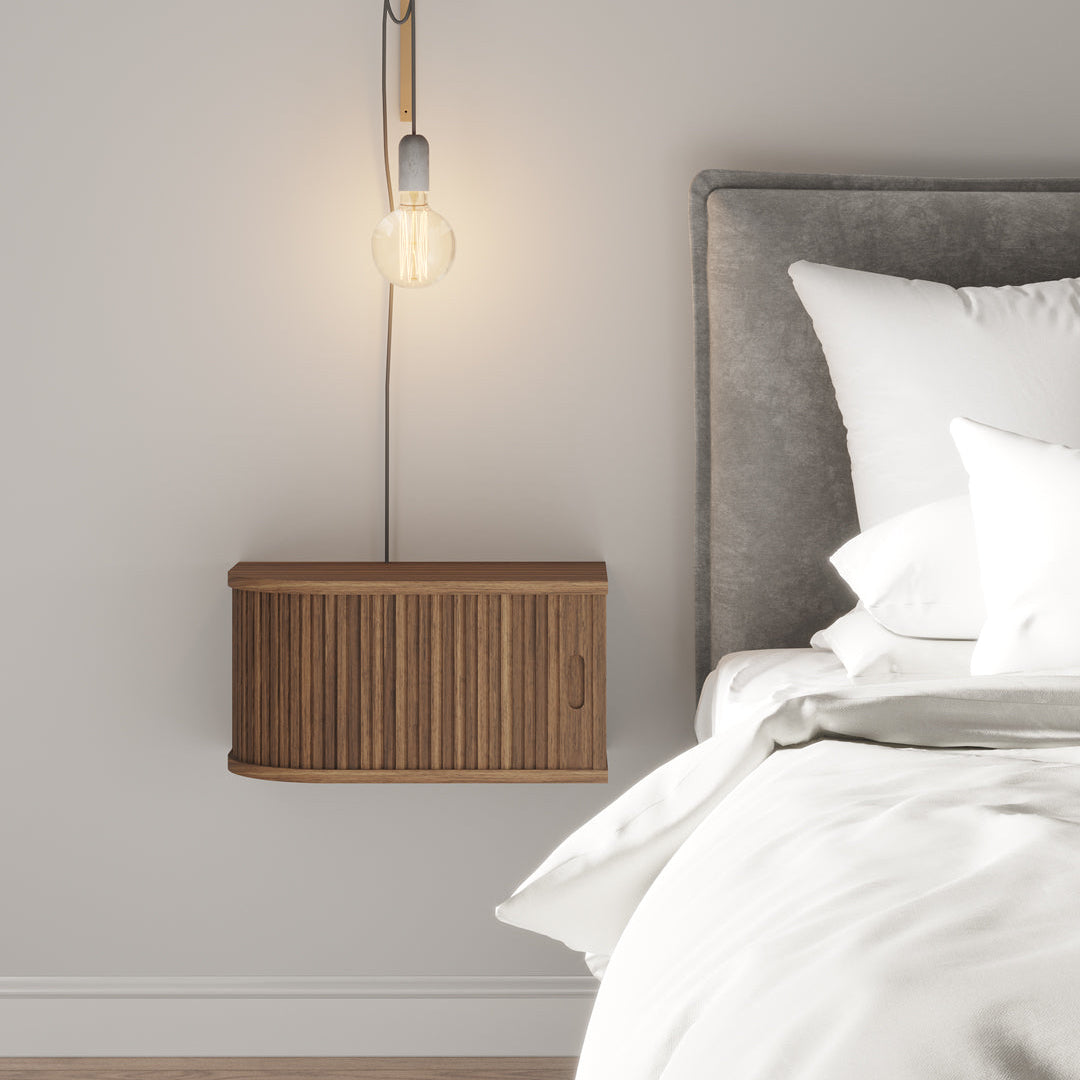 A modern wall-mounted bedside lamp with a warm glowing bulb, featuring a curved brass-toned metal top and a ribbed wooden cabinet base, shown next to a bed with white bedding and a grey headboard.