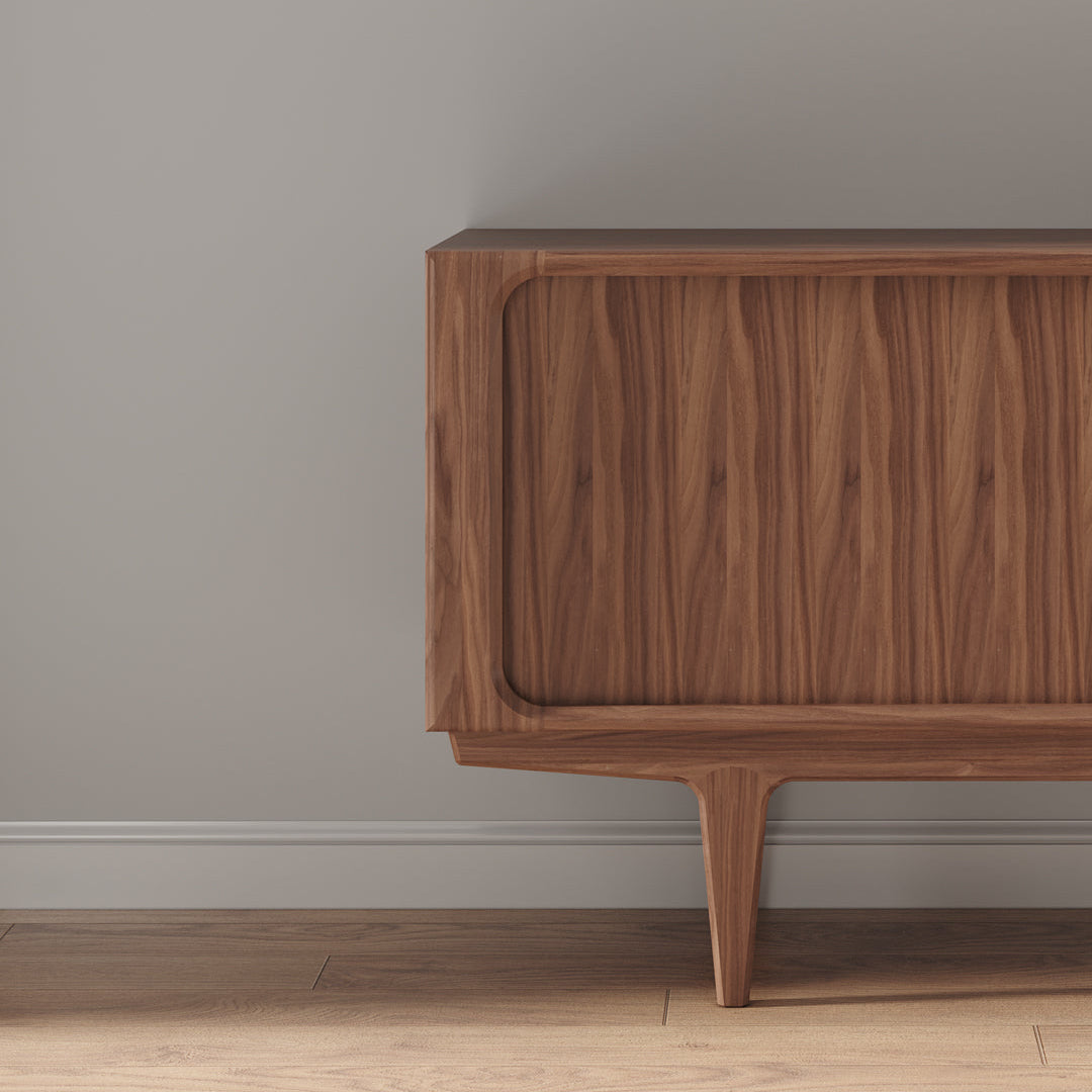 A close-up of a mid-century modern wooden sideboard or credenza with a warm walnut finish, set against a neutral gray wall and light wood floor.