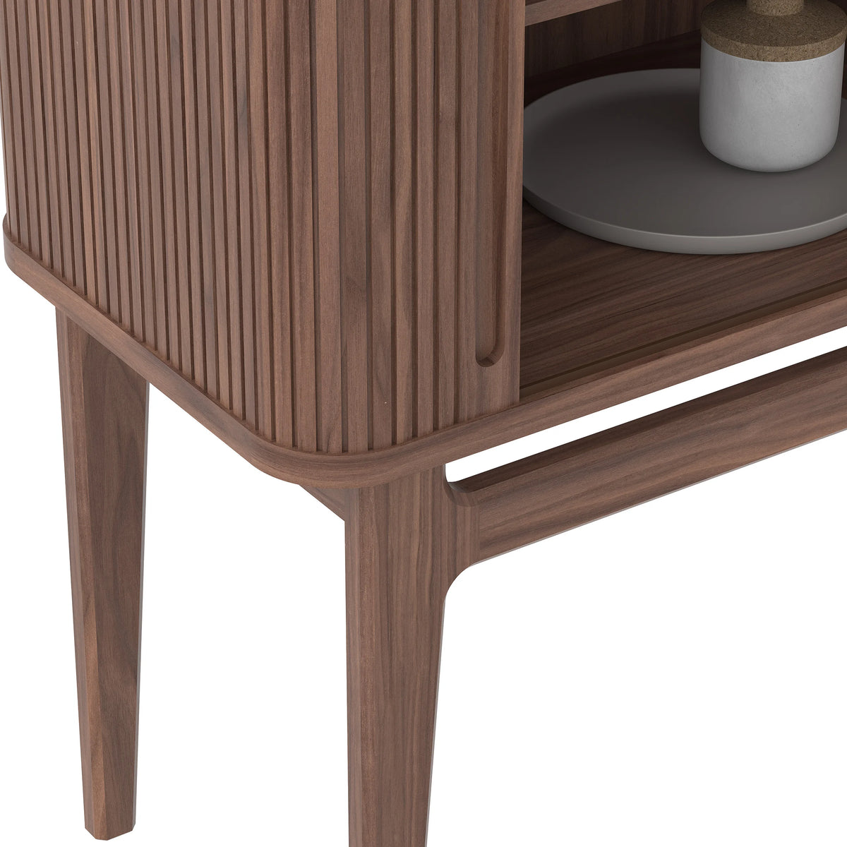 Close-up of a wooden cabinet  with a slatted front panel and tapered legs, shown against a white background with a small tabletop decor item partially visible.