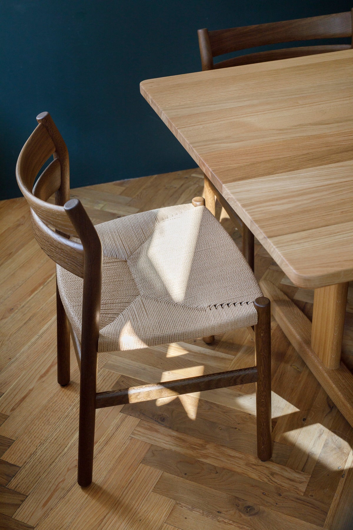A modern wooden dining chair with a woven beige seat, positioned next to a matching wooden table on a herringbone wood floor with natural sunlight casting shadows.