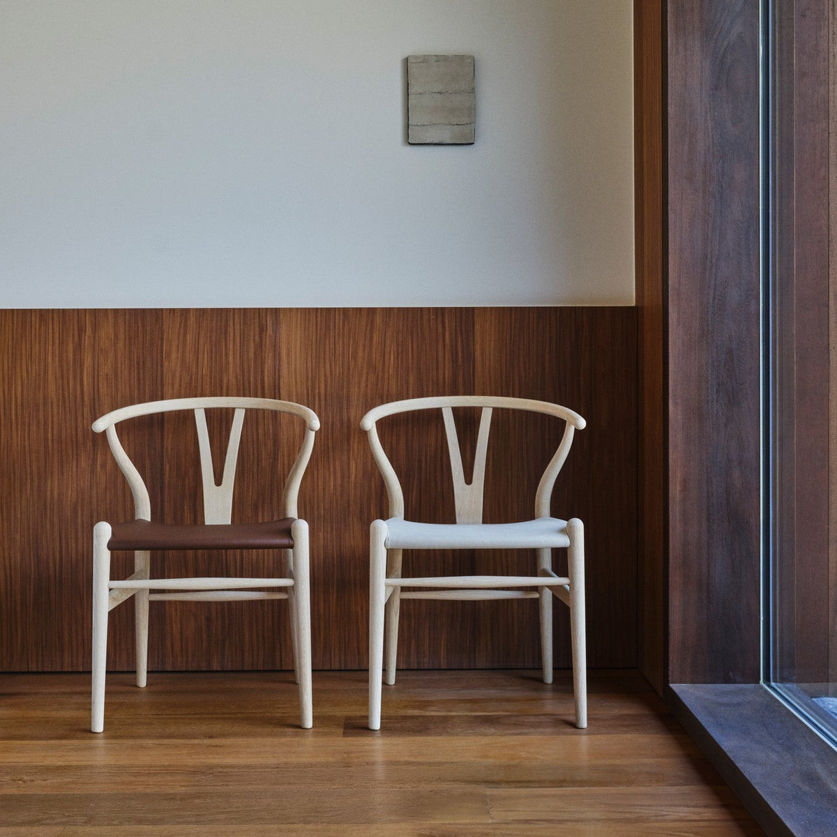 Two white Wishbone chairs with dark brown woven seats, positioned side-by-side against a wood-paneled wall and hardwood floor.