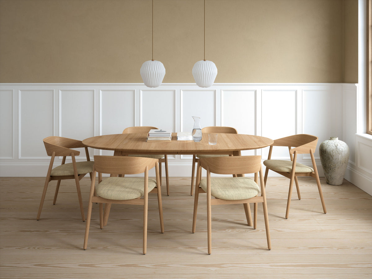 A modern dining set featuring a round light oak wood table and six matching chairs with light beige upholstered seats, arranged in a bright room with a beige and white paneled wall, hanging pendant lights, and a large ceramic vase.