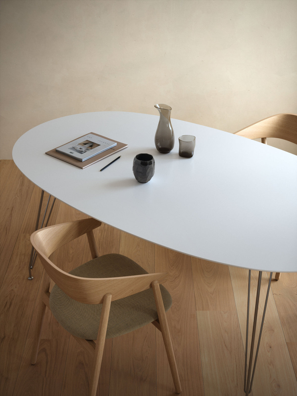 An oval white dining table with thin metal hairpin legs, styled with light wood chairs, a ceramic pitcher, and a small dark cup on a light wood floor against a beige wall.