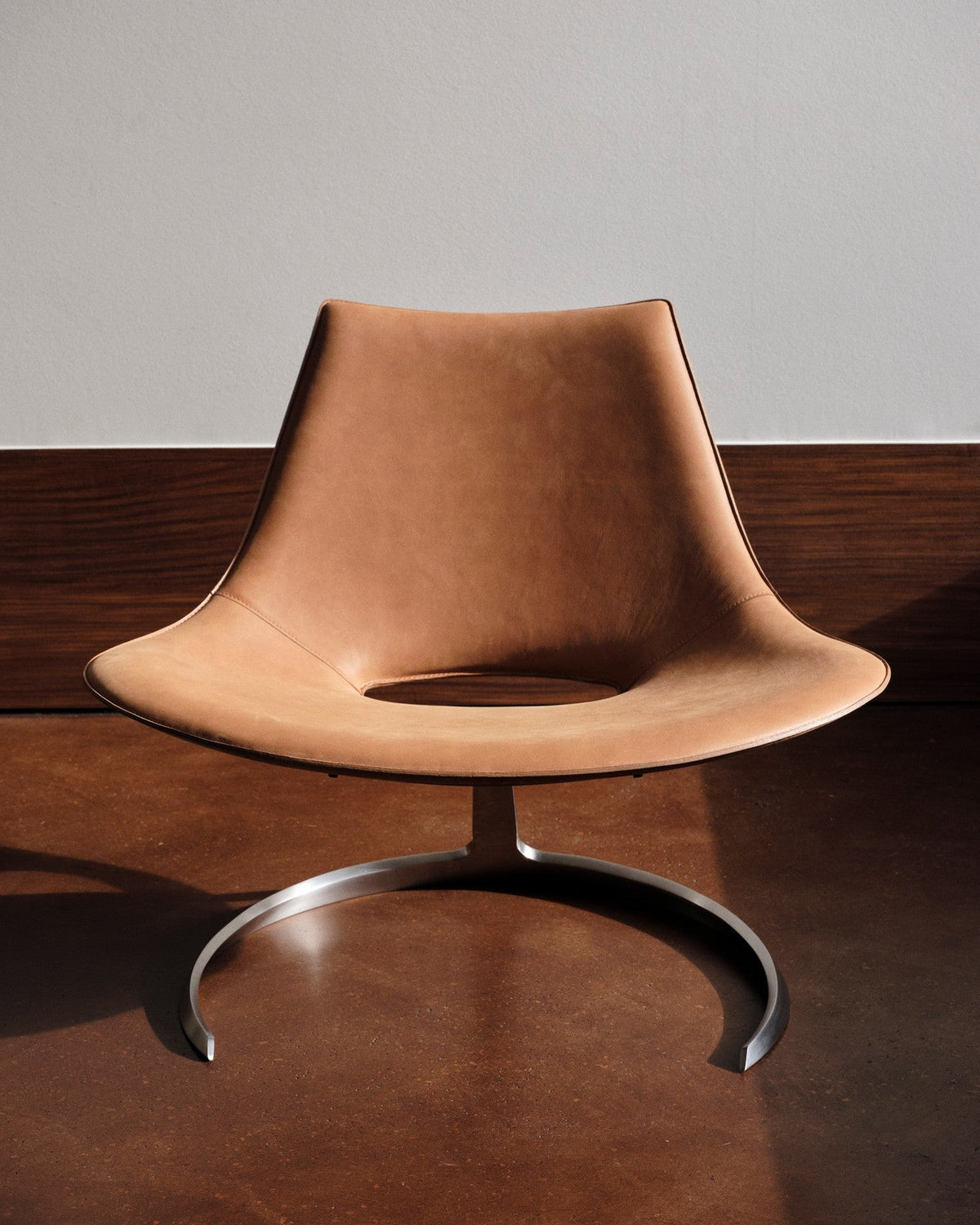 A modern sculptural leather chair with a curved tan leather seat and backrest, supported by a polished silver metal base, set against a dark wood panel and light gray wall background.