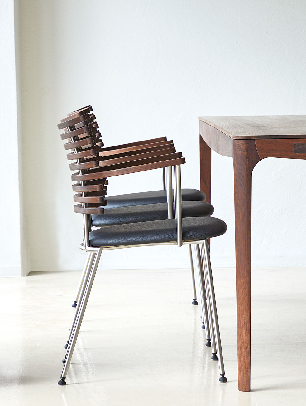 Three modern dining chairs with dark brown wooden slatted backs and seats with black cushions, arranged next to a wooden table in a minimalist room.