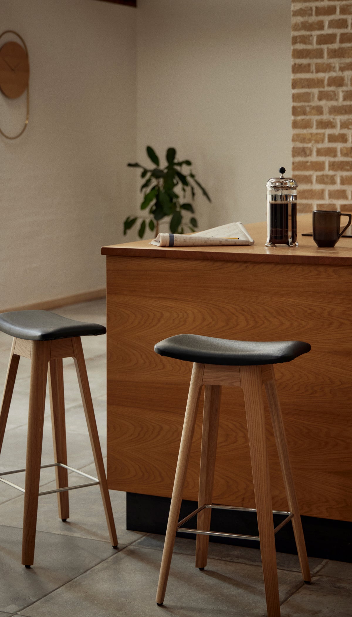 Two wooden bar stools with black seat cushions in a modern kitchen or cafe setting, with a wood counter, coffee press, and brick wall in the background.
