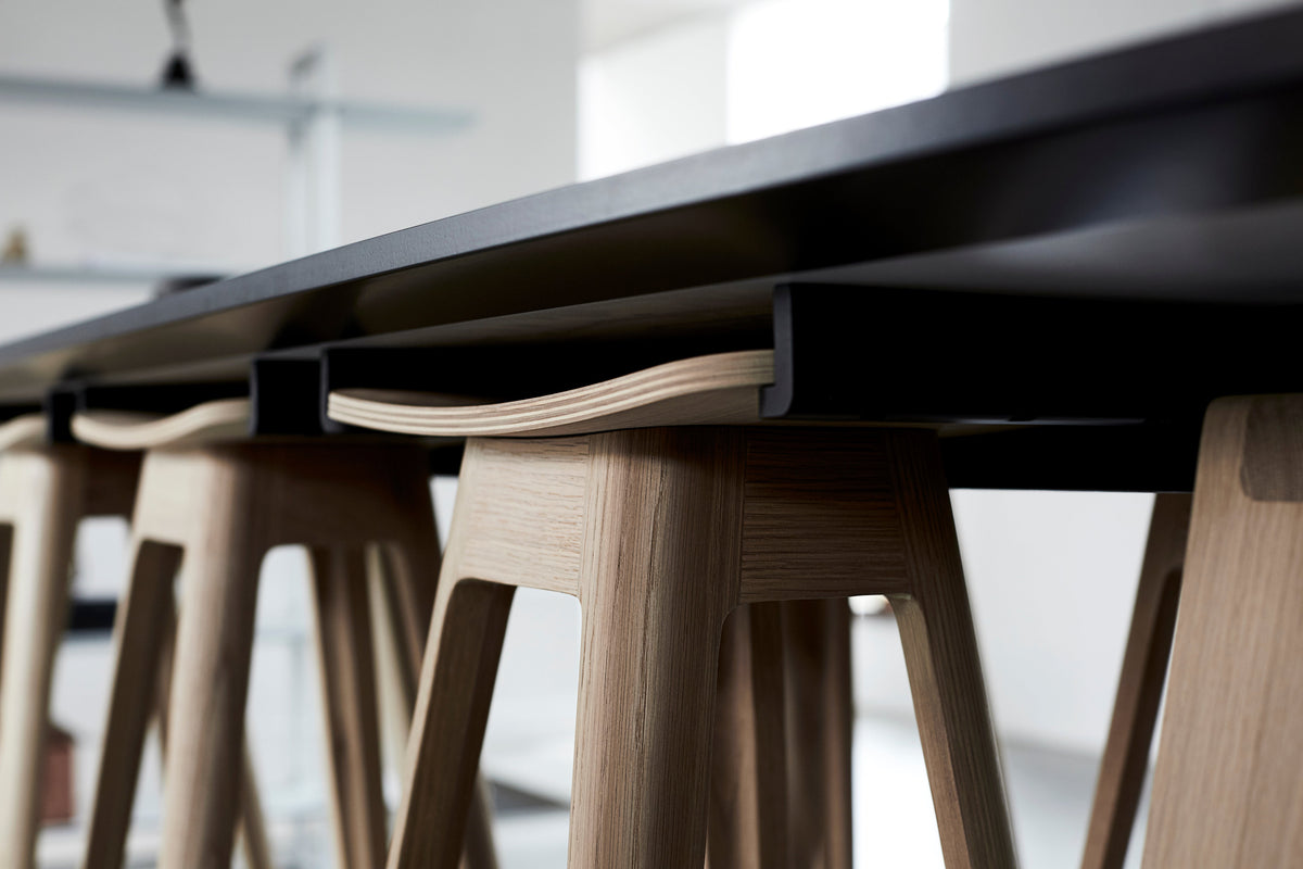 Close-up of a modern black bar table with  light natural wood bar stools hanging on brackets to the underside of the table shown in a bright interior setting.