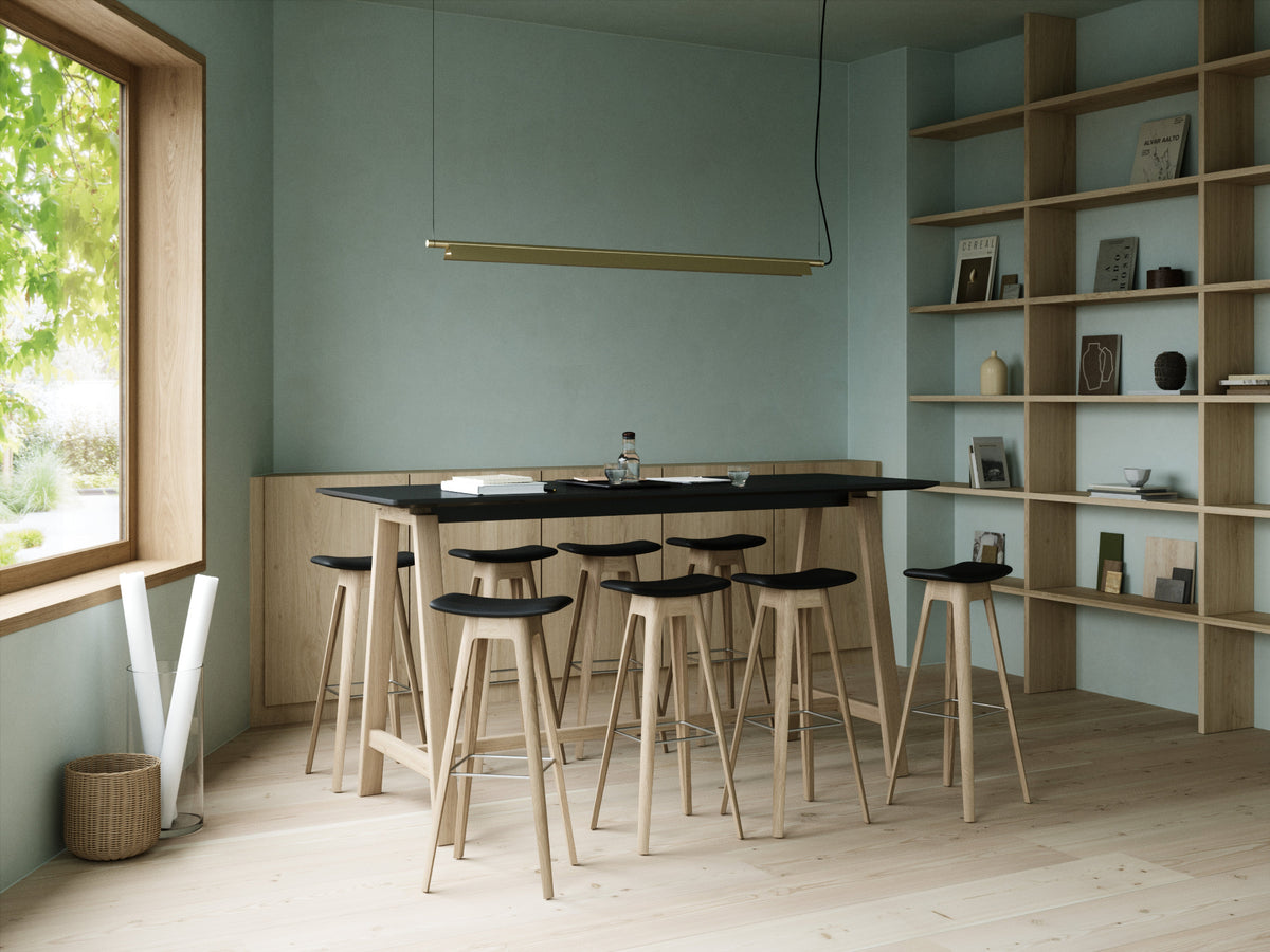 A modern dining area featuring a tall wooden bar-height table with a black top and eight matching wooden bar stools with black seats, set against a sage green wall with built-in shelving.