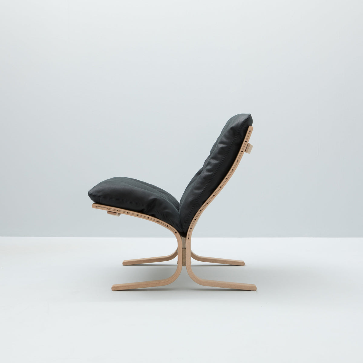 A modern lounge chair with black leather upholstery and a light natural wood frame, shown from the side against a minimalist light gray background.