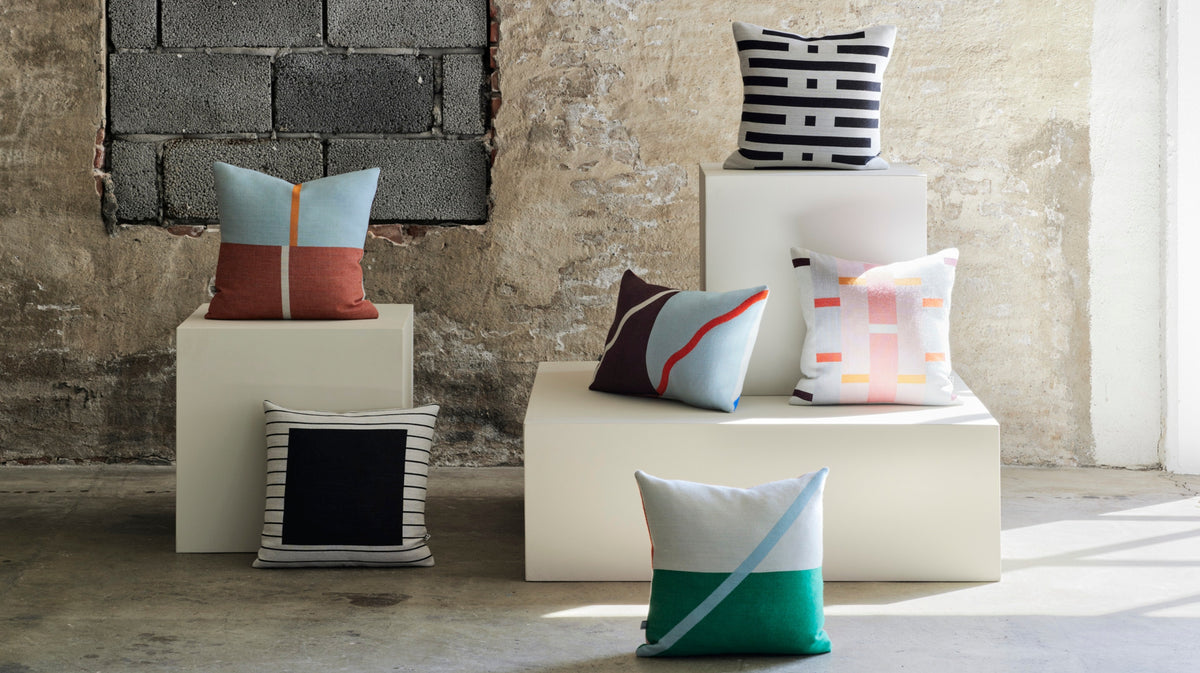 A collection of colorful geometric throw pillows displayed on white display blocks in a rustic, sunlit room with a concrete floor and exposed brick wall.