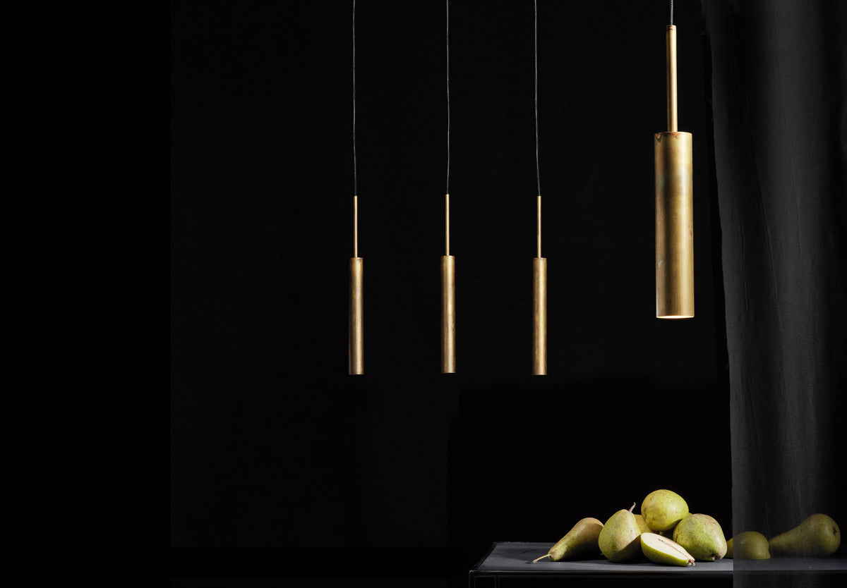 Three modern cylindrical pendant lights in a raw brass hang against a dark black background, with a small table at the bottom holding green pears.
