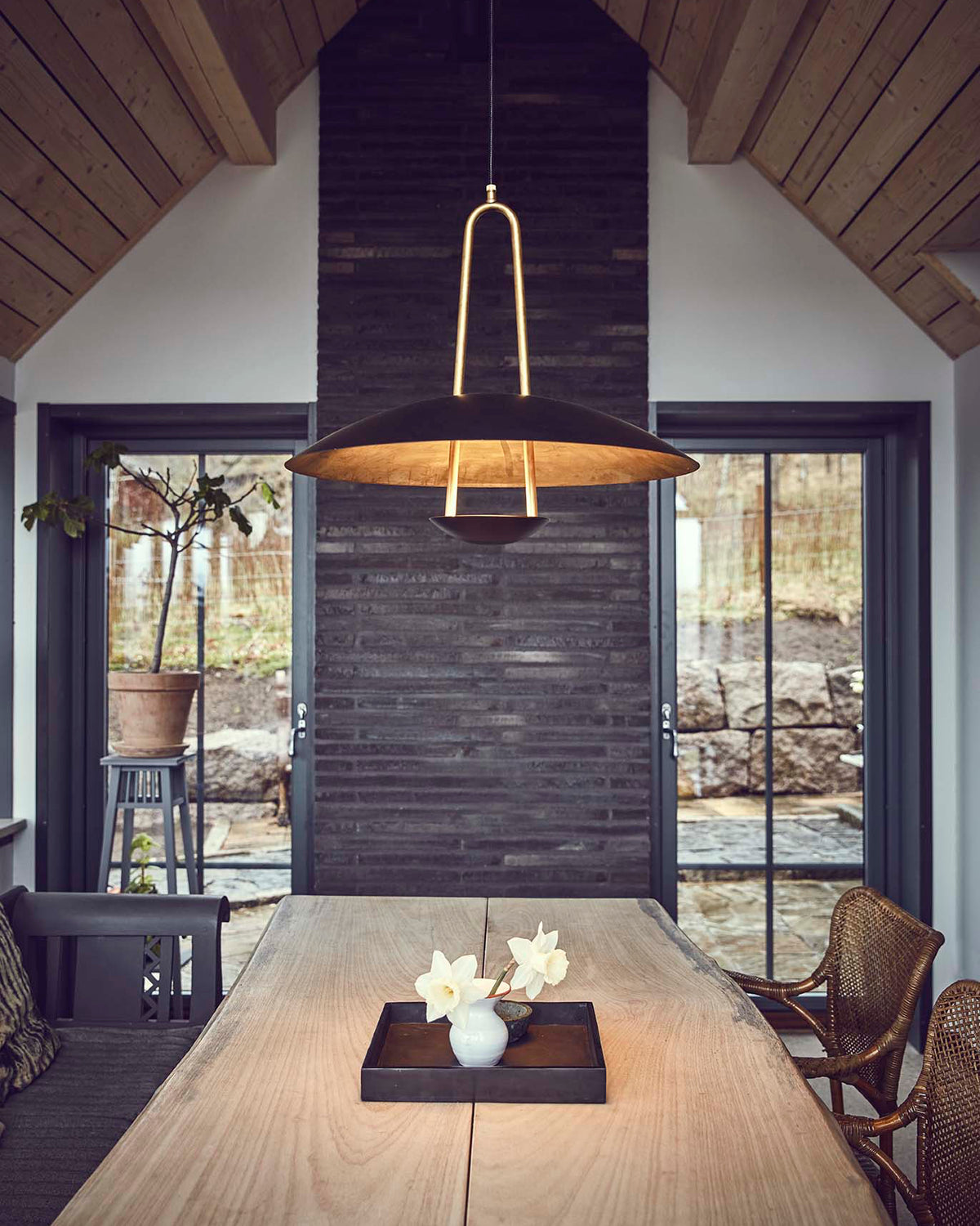 A modern pendant light with a dark bronze conical shade and a gold-toned U-shaped top bar hangs over a long wooden dining table in a rustic cabin interior.