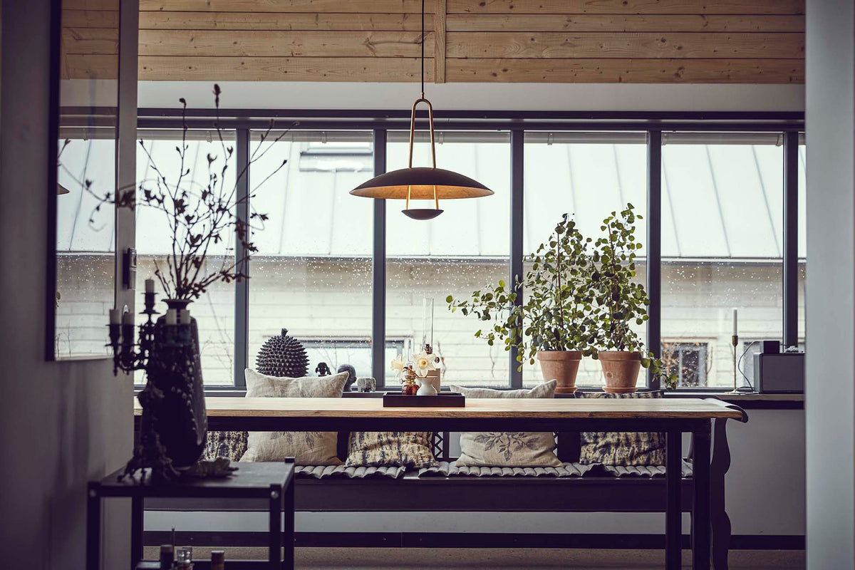 A modern pendant light with a bronze-toned metal frame and a warm glowing dome shade hangs over a wooden dining table in a bright, contemporary room with large windows and plants.