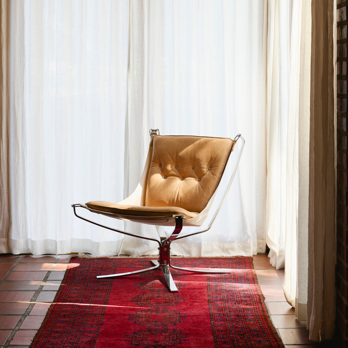 A mid-century modern lounge chair with tan leather upholstery and a chrome base, positioned on a red patterned rug in front of sheer white curtains.