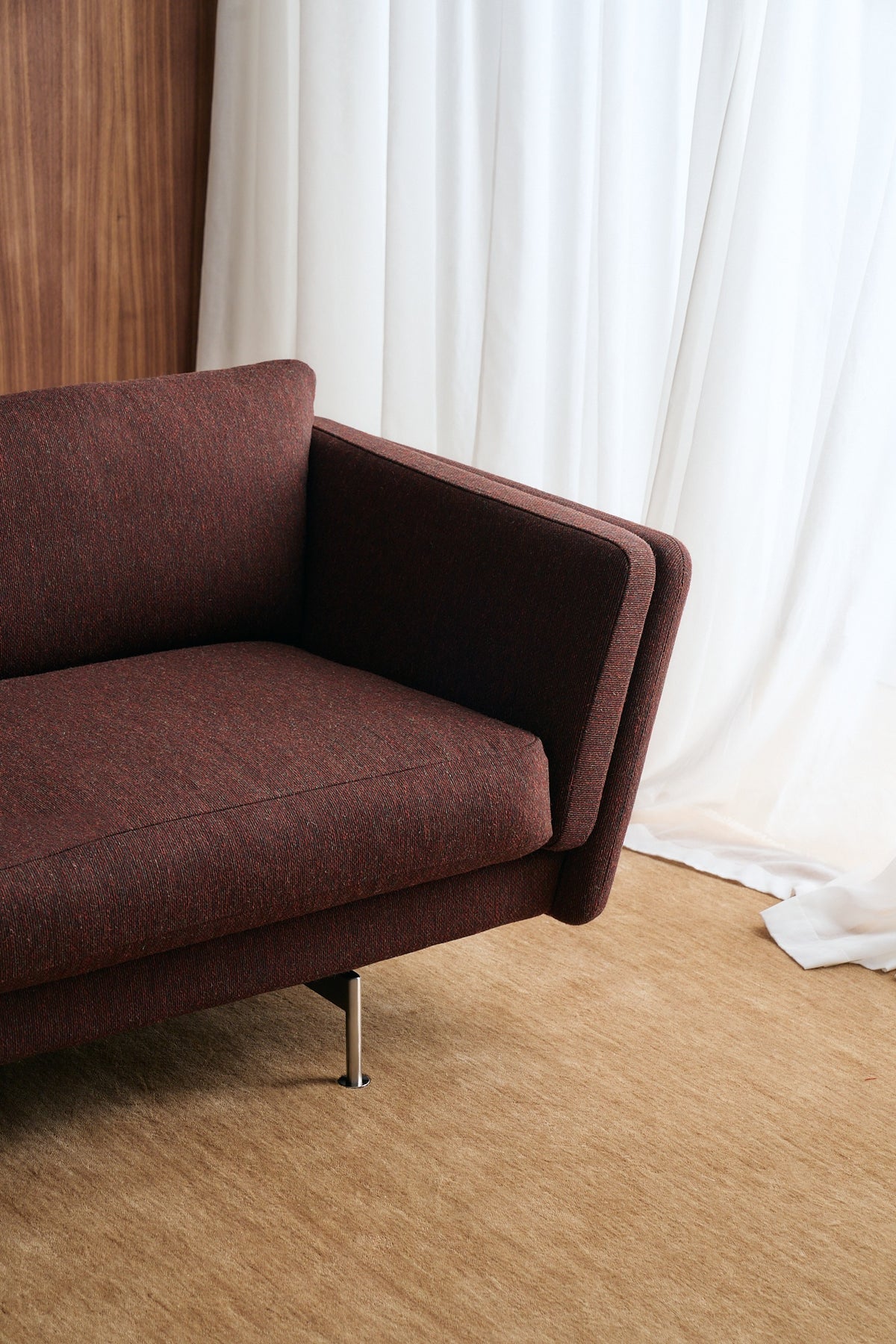 A close-up of a modern sofa in a rich dark brown fabric, positioned on a beige carpet in front of white curtains.
