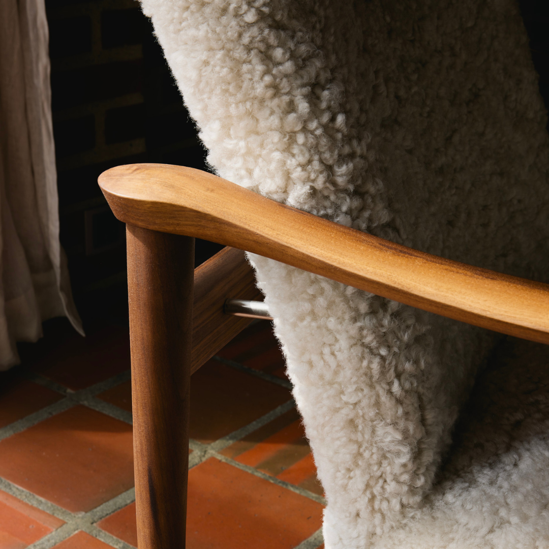 Close-up of a wooden armchair with a thick, fluffy off-white sheepskin or wool seat and back, set in a room with terracotta tile flooring and a curtain in the background.