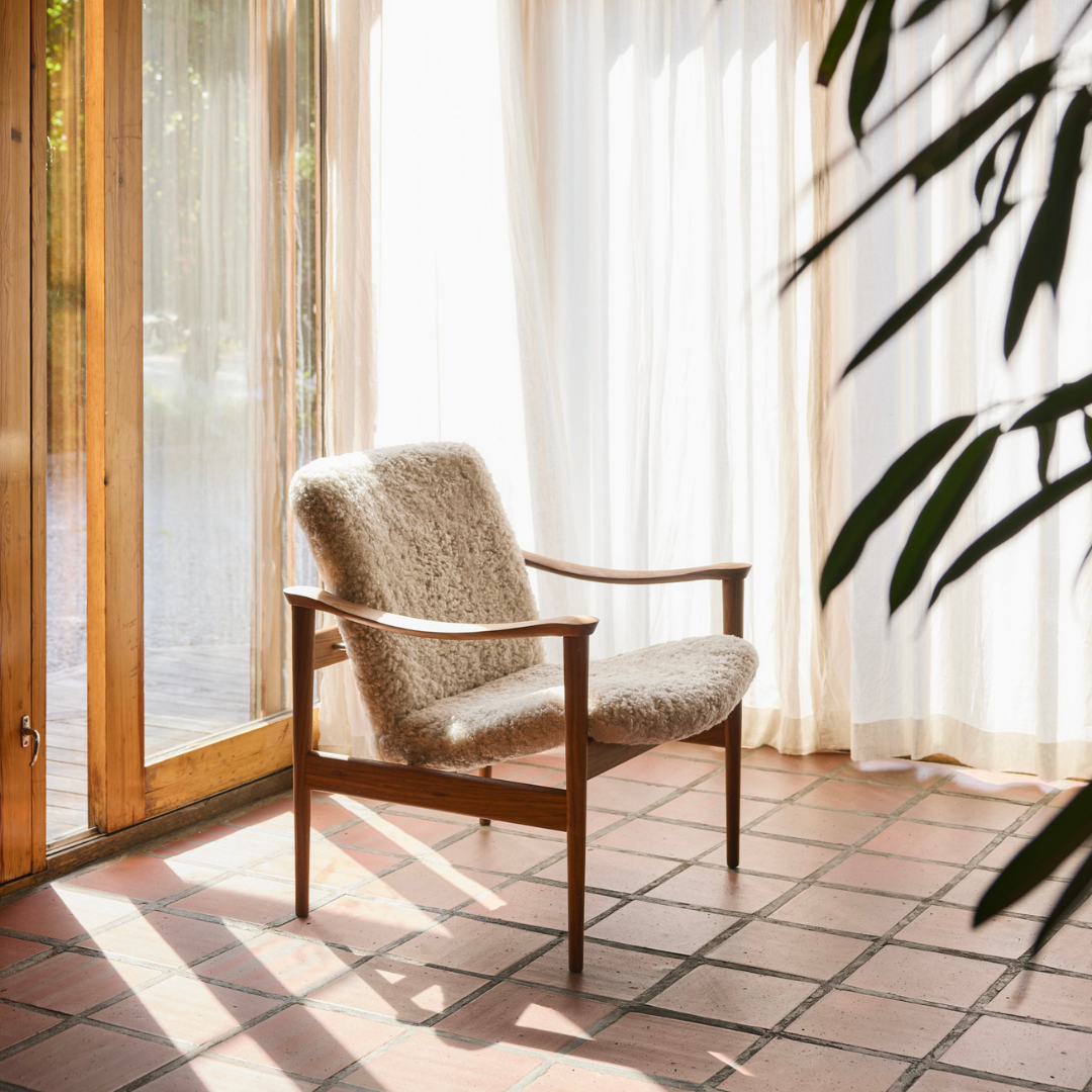 A mid-century modern armchair with a textured beige bouclé seat and backrest and a warm brown wooden frame, positioned on a terracotta tiled floor in a sunlit room with sheer white curtains and a glass door.