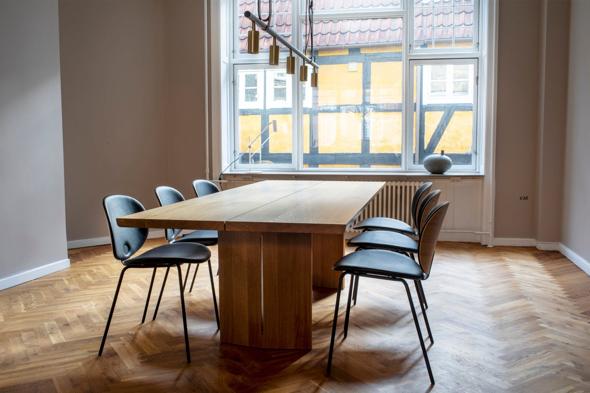 A modern dining room setup featuring a rectangular wooden dining table with a thick pedestal base, surrounded by six black chairs with gray-blue seats and black metal legs. The room has light beige walls, a herringbone wood parquet floor, and a large multi-pane window looking out onto other buildings.