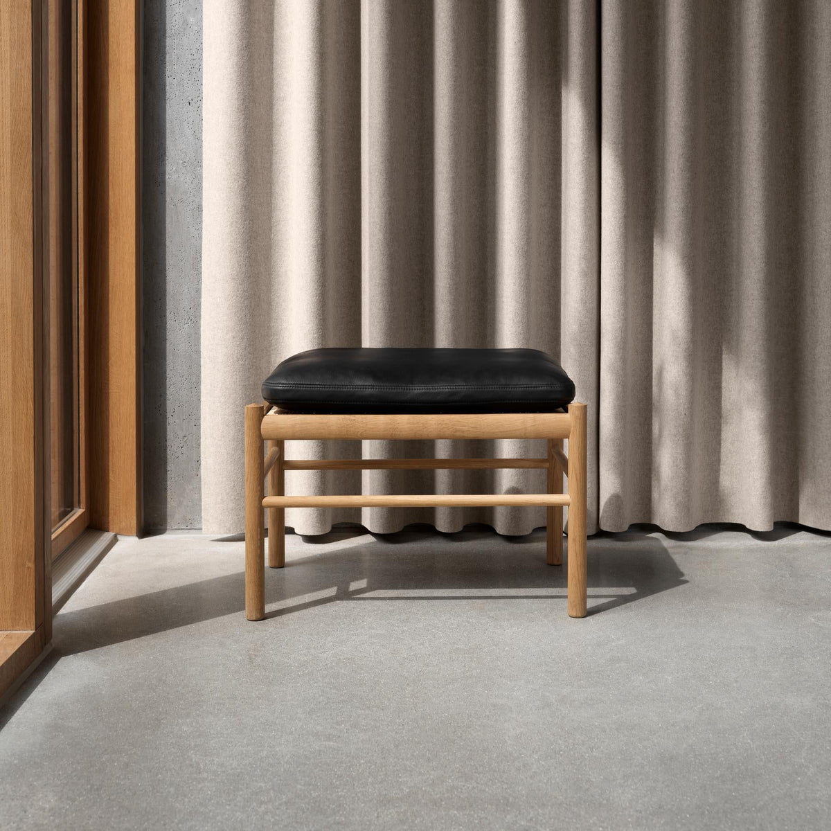 A minimalist wooden stool with a black cushioned seat, positioned in front of a floor-length beige curtain with soft vertical shadows.