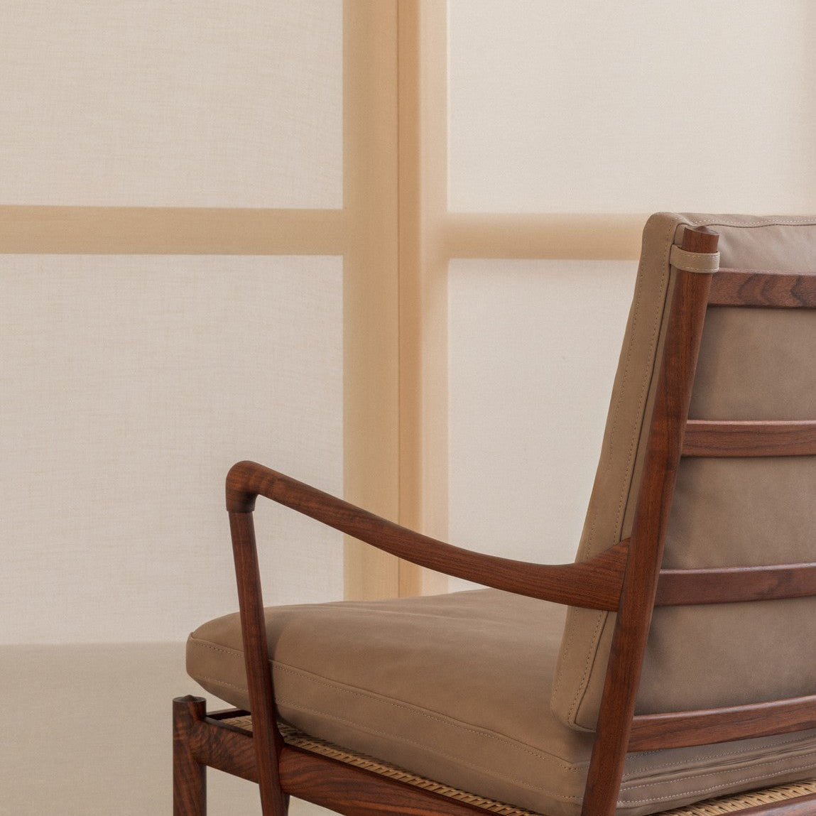 A close-up of a mid-century modern armchair with a warm brown wooden frame and beige cushioned seat and backrest, set against a minimalist cream-colored wall with a vertical wooden divider.