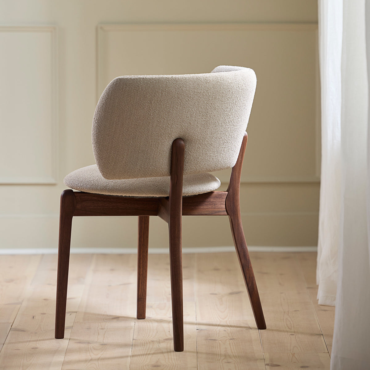 A mid-century modern dining chair with a cream bouclé upholstered back and seat, supported by a dark walnut wood frame, set in a minimalist room with beige walls and light wood floors.