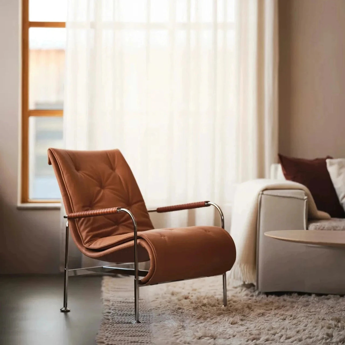 A mid-century modern lounge chair upholstered in warm cognac brown leather with a chrome metal frame, positioned in a stylishly decorated living room next to a window with sheer curtains.