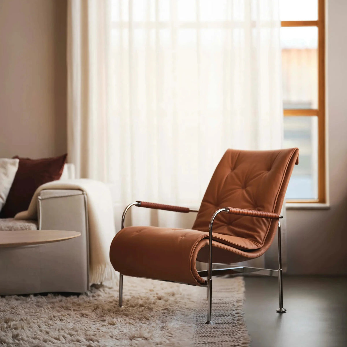 A mid-century modern accent chair upholstered in warm brown tufted fabric with a curved chrome metal frame, positioned in a stylishly decorated living room next to a large window with sheer white curtains.