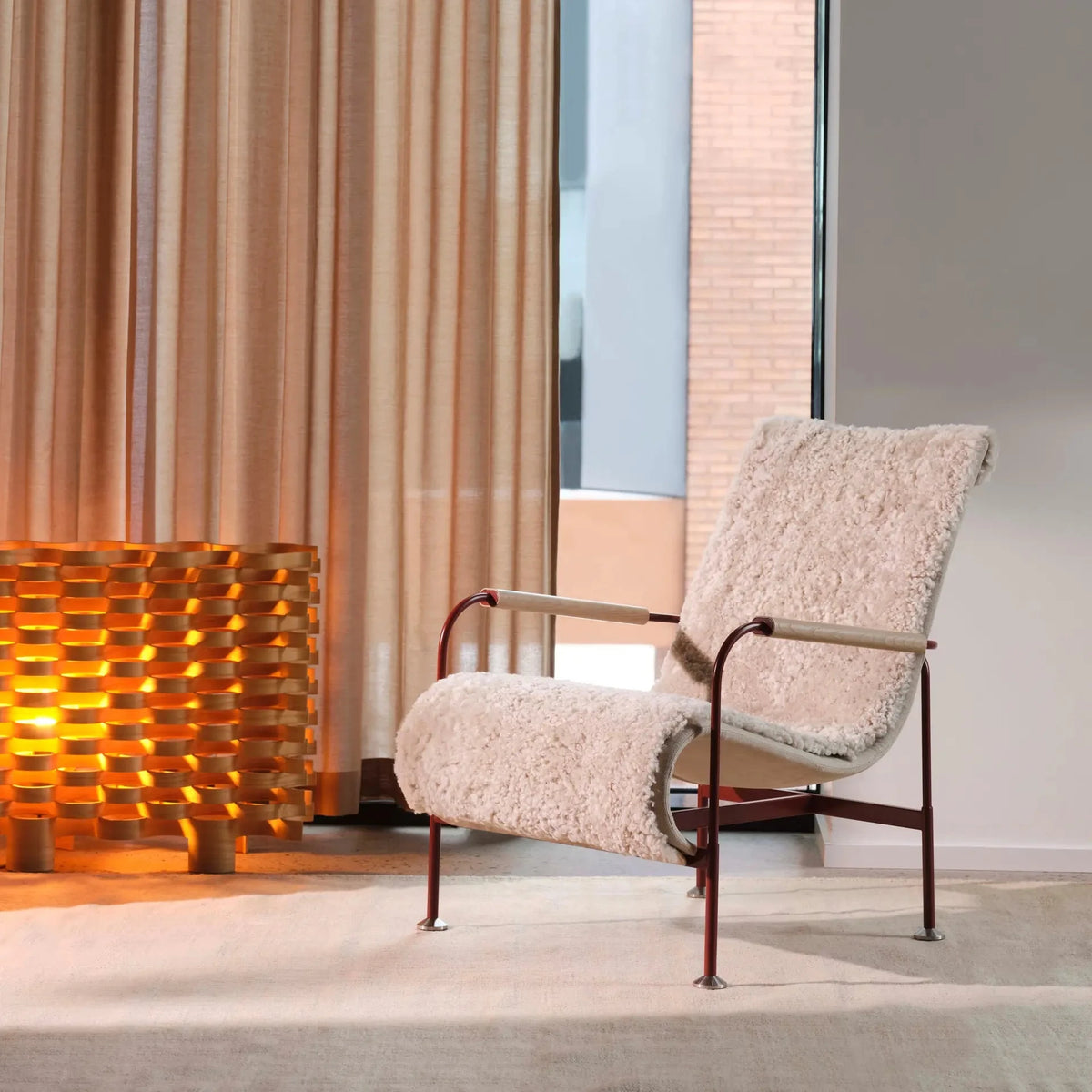A modern lounge chair with textured cream bouclé upholstery and a dark metal frame, positioned in a minimalist interior setting next to a warm glowing woven floor lamp and beige curtains.