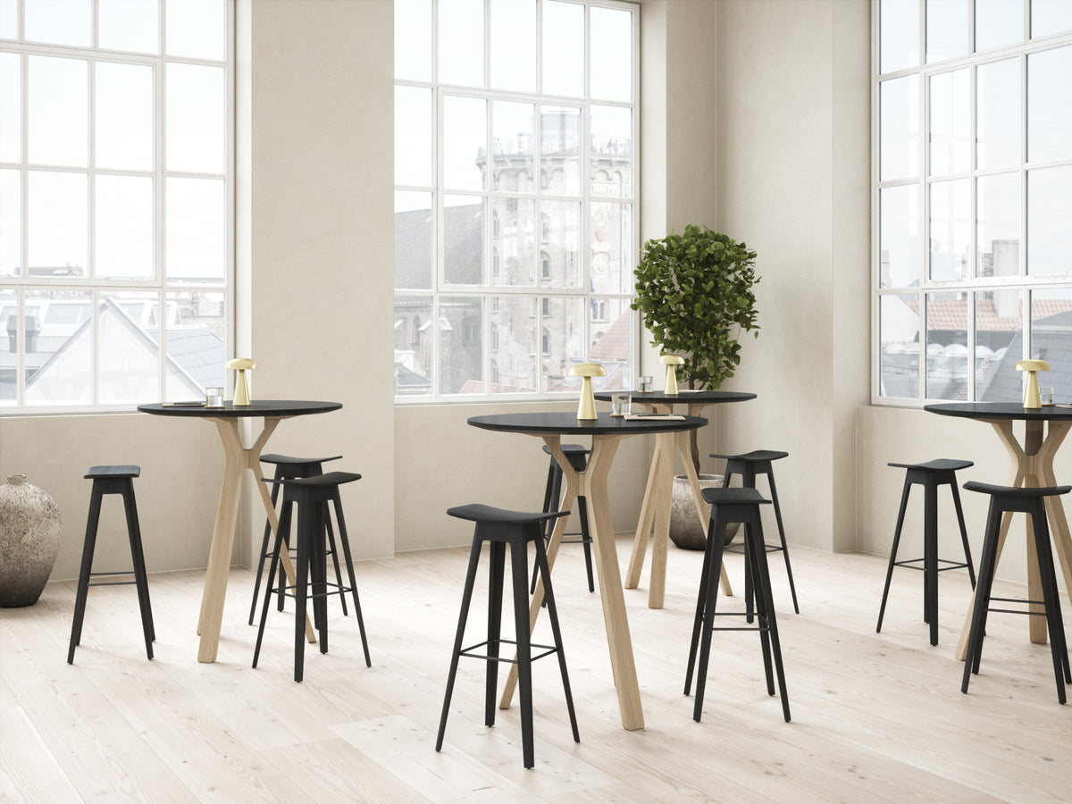 A modern dining or bar-height table set with round black tops and light wood legs, paired with black stools, arranged in a bright, minimalist room with large windows and light wood flooring.
