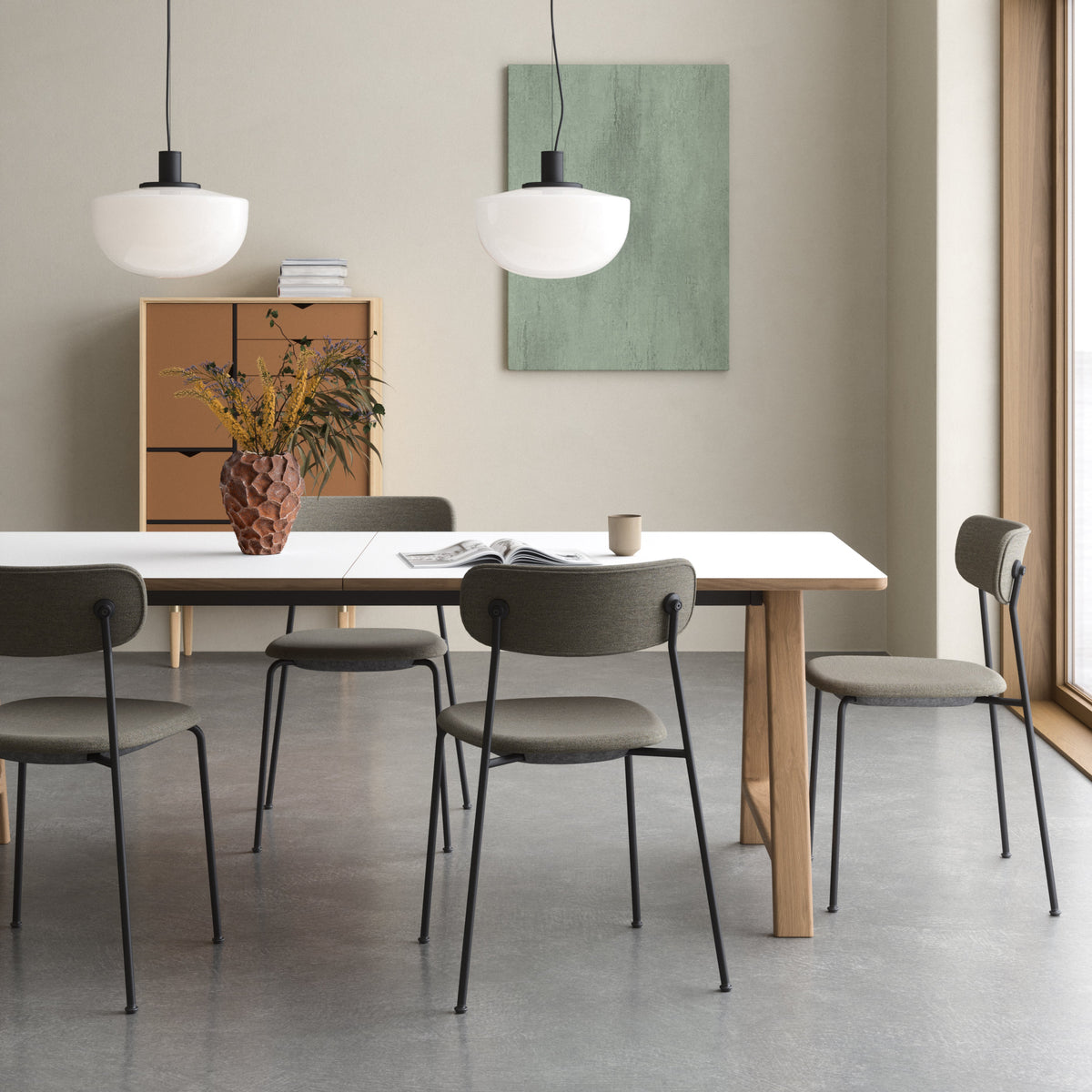 A modern dining room scene featuring a white rectangular table with four gray upholstered chairs, two white pendant lights, a wooden side cabinet, and a green wall art piece.