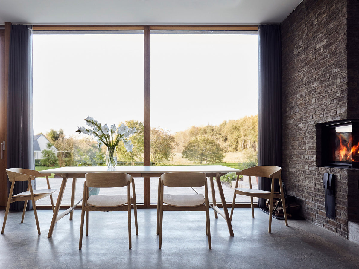 A modern dining room scene with a light wood dining table and four matching wooden chairs, positioned in front of large floor-to-ceiling windows. The room features a dark brick wall with a built-in fireplace on the right side, and an outdoor view of trees and grass.