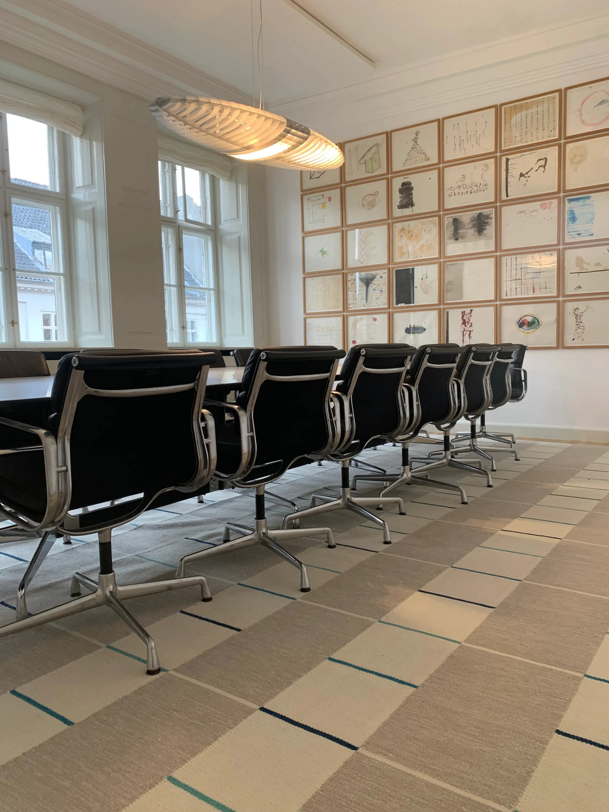 A long conference table surrounded by black office chairs with chrome bases in a bright, modern meeting room. The room has large windows, a patterned beige carpet, and a wall covered with framed artwork.