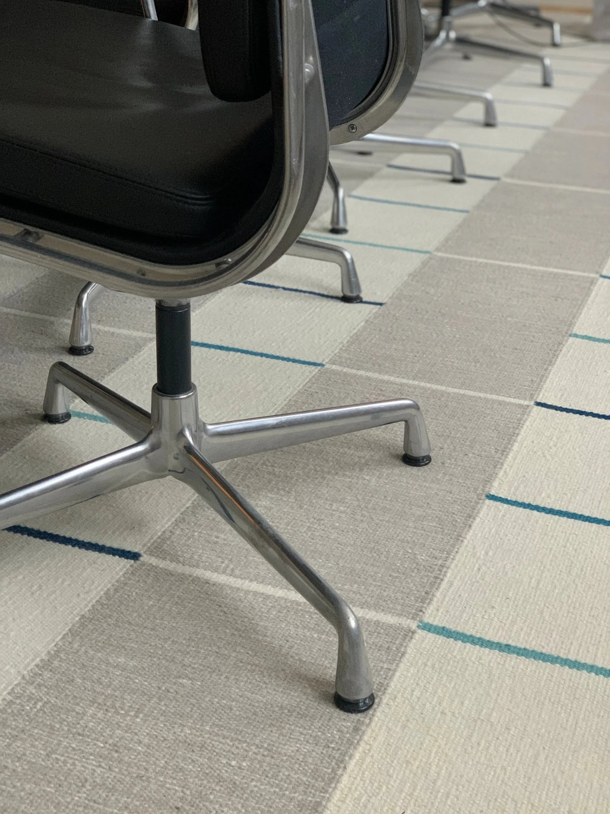 A close-up, low-angle view of a modern office chair with a black seat and a chrome five-star base, positioned over a textured beige and gray commercial carpet with thin blue stripes.