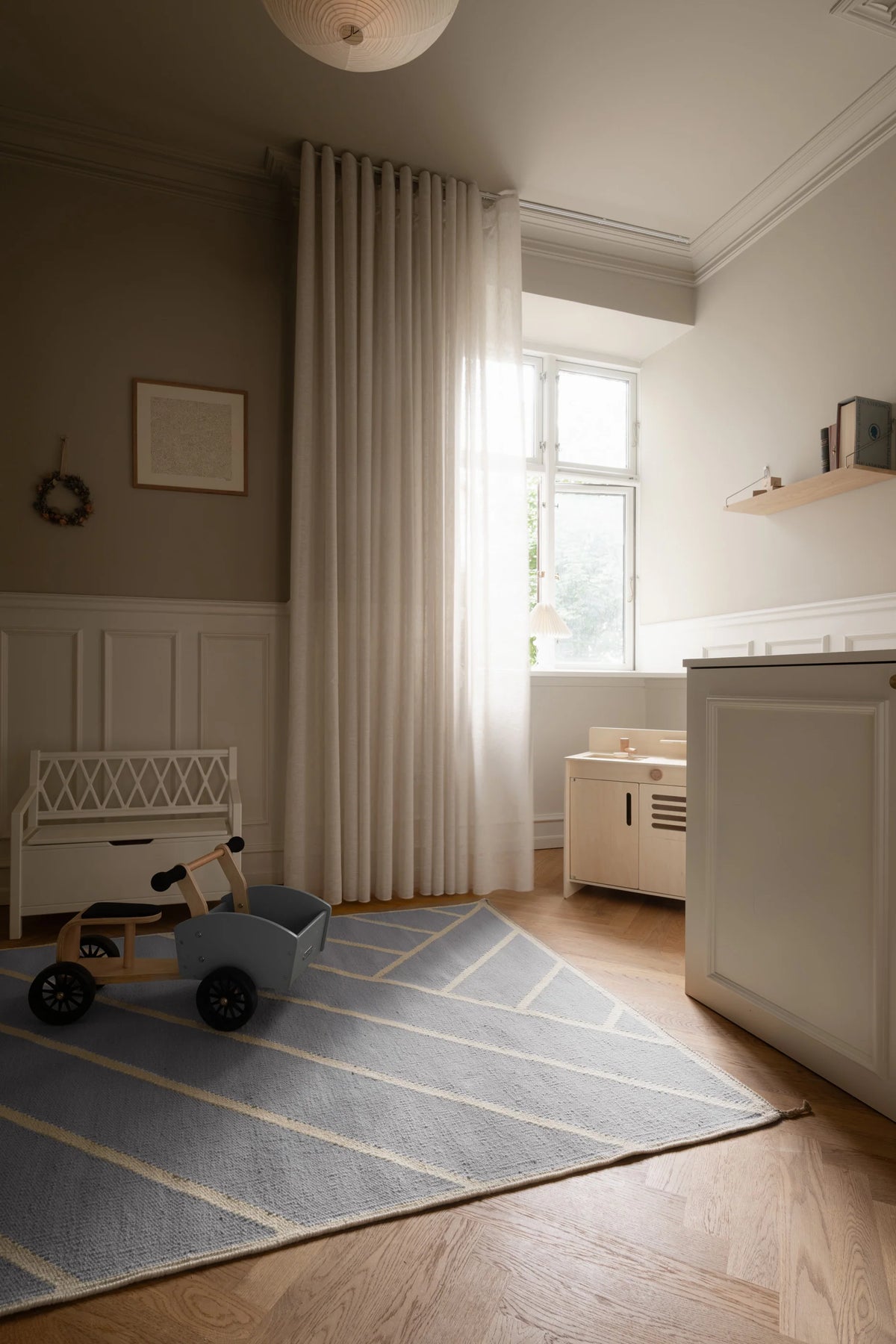 A styled children's room corner featuring a light blue and beige geometric patterned area rug on a wooden floor, with a white bench, wooden toy car, and a tall cream pleated curtain hanging by a window.
