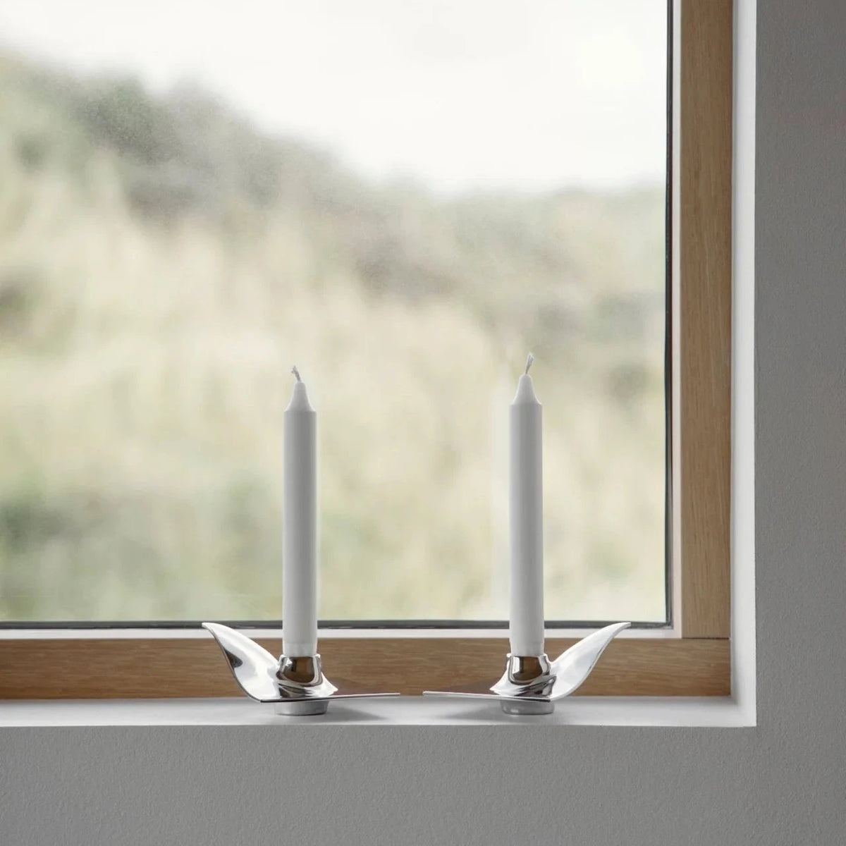 Two white candles in silver candle holders on a windowsill with a blurred natural background